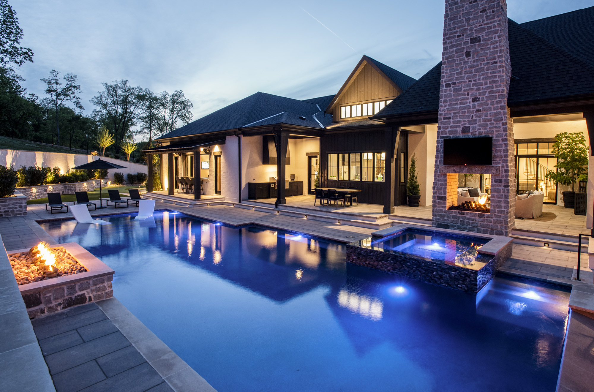Luxurious modern home exterior at dusk featuring an illuminated swimming pool, integrated spa, fire pit, and contemporary outdoor living spaces.