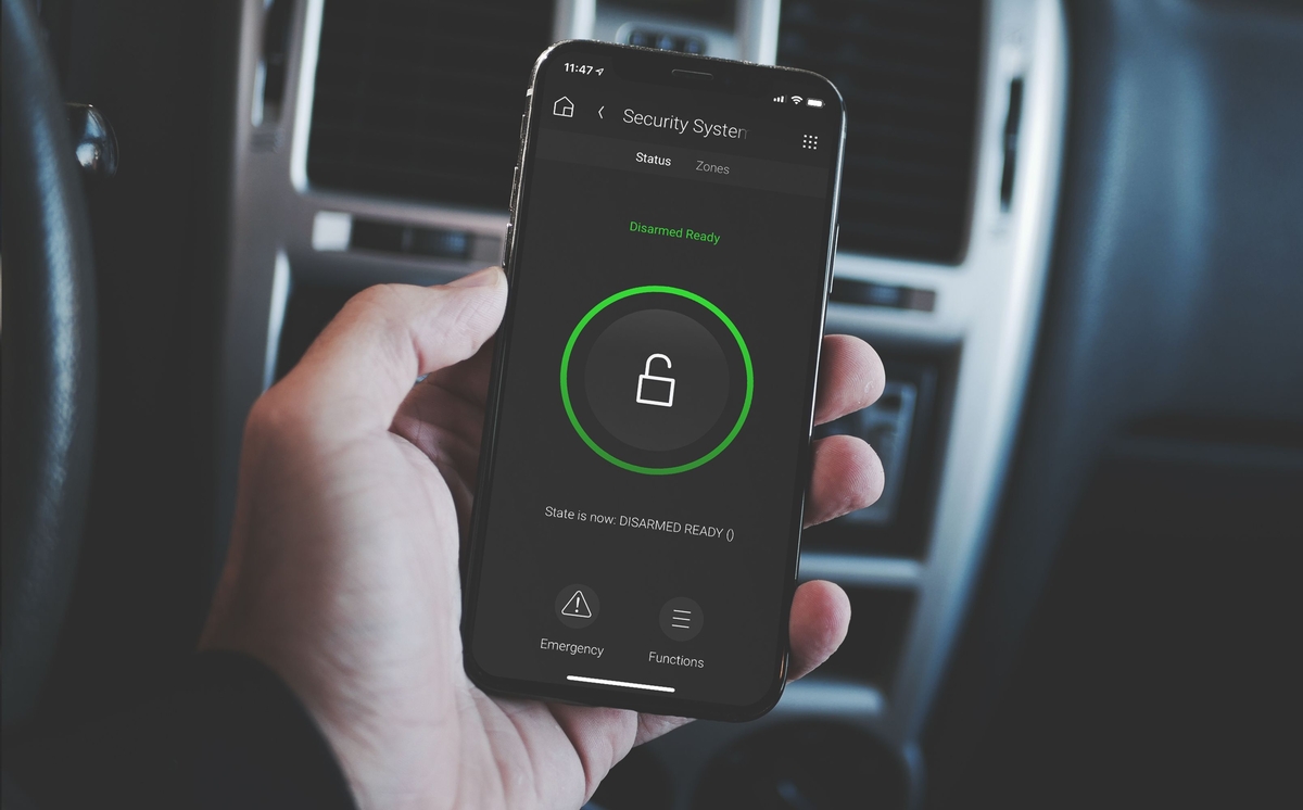 Person holding a smartphone inside a vehicle displaying a security app screen showing a green unlocked padlock and disarmed system status.