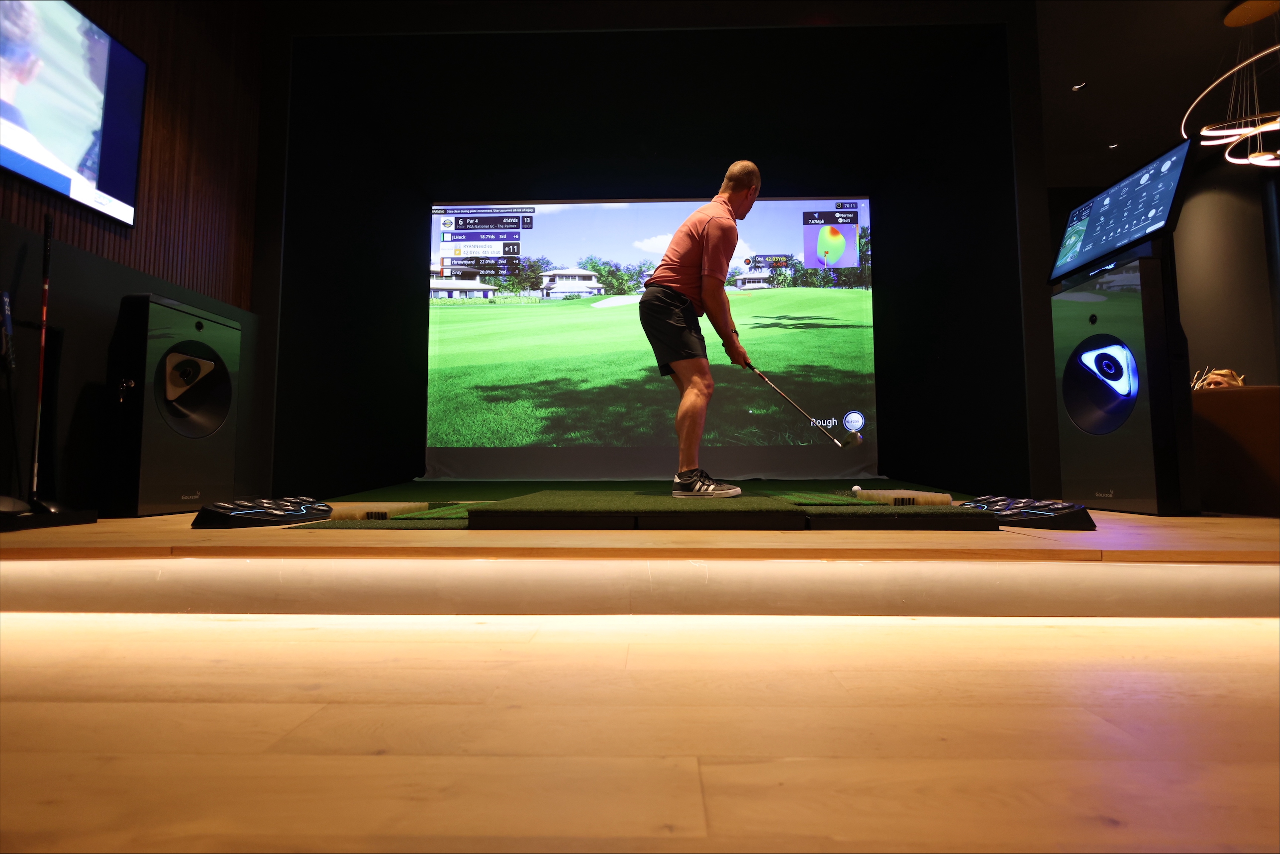 Golfer practicing a swing in an indoor golf simulator bay, standing on a hitting mat facing a large projected virtual golf course.
