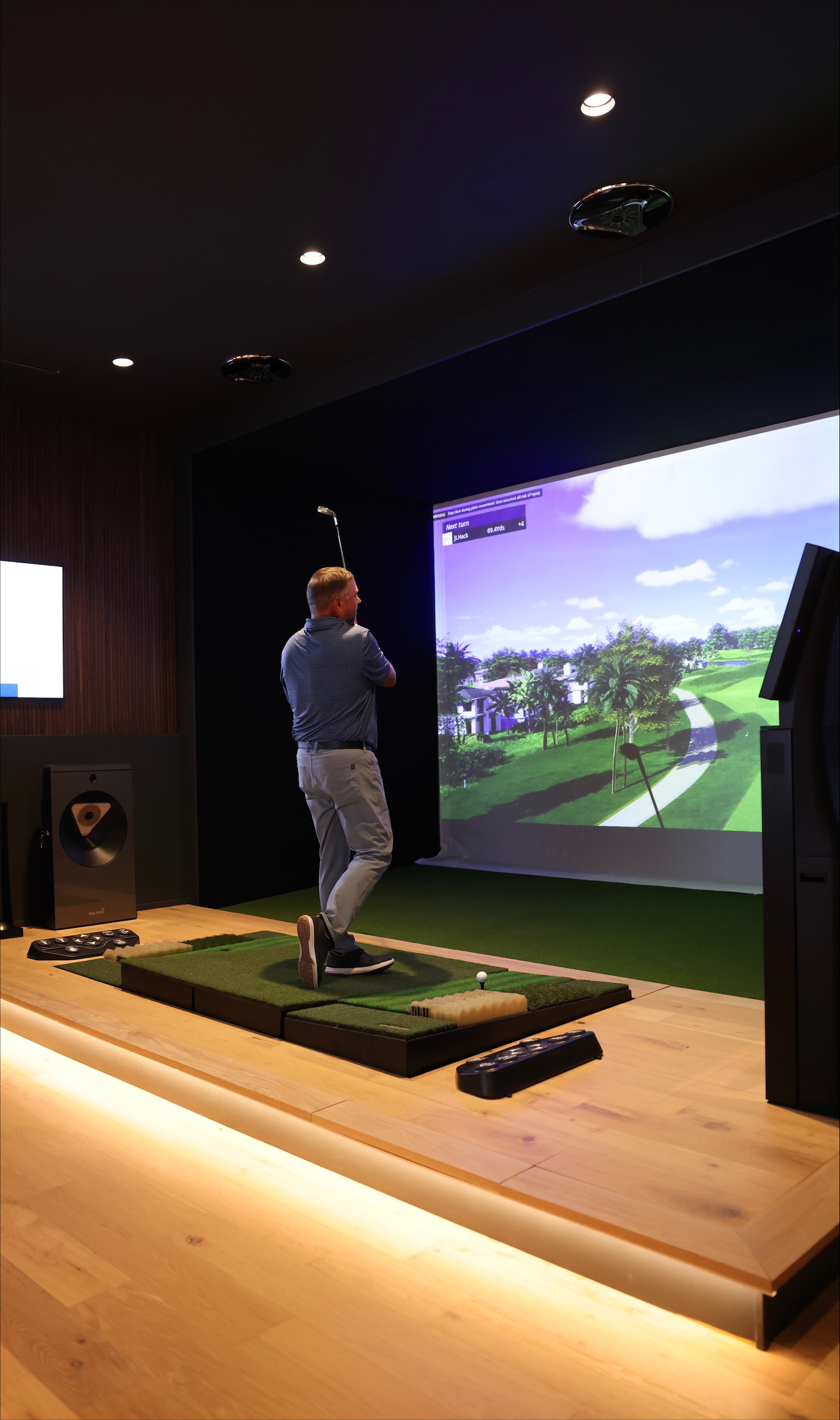 Golfer finishing a shot in an indoor golf simulator, with a projected fairway on a large screen and a ball positioned on the hitting mat.