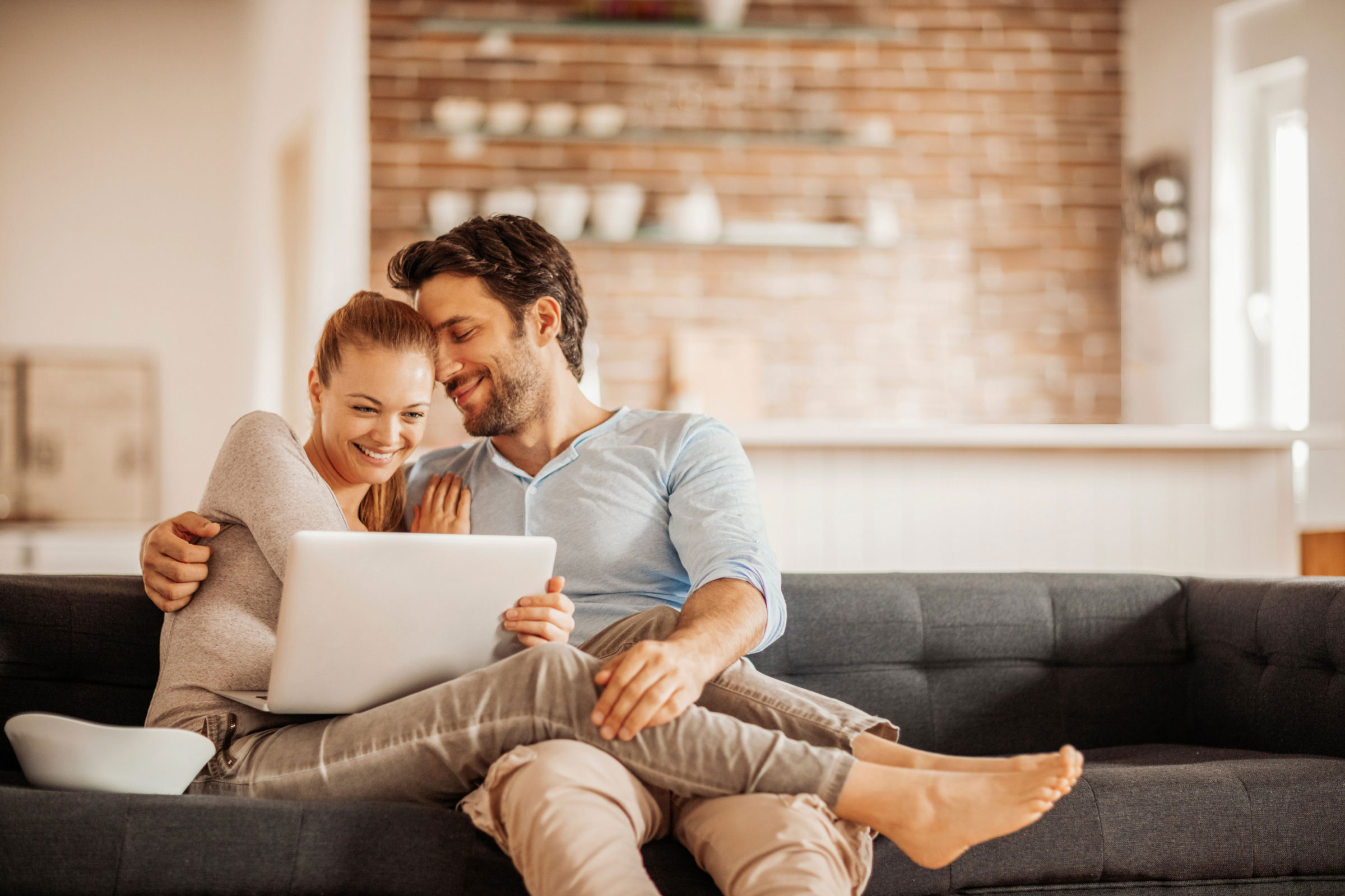 Two people sitting closely on a dark gray couch looking at a white laptop together in a cozy modern living room with a brick wall and shelves in the background.