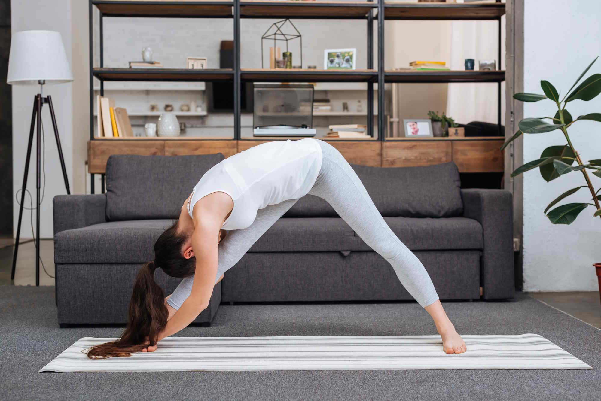Person practicing a forward bend yoga pose on a striped mat in a cozy living room with a gray sofa, shelving unit, and floor lamp.