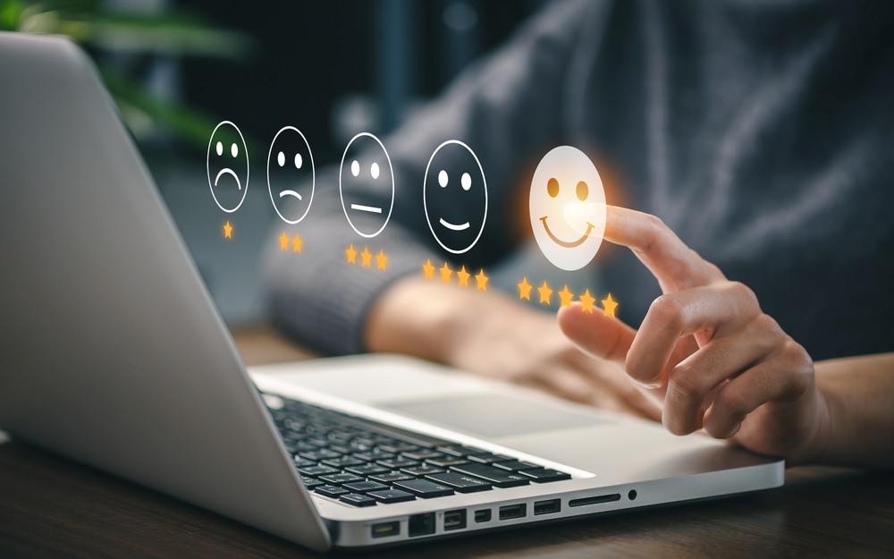 A person selects a five-star smiley face rating on a virtual feedback scale displayed above a laptop keyboard.