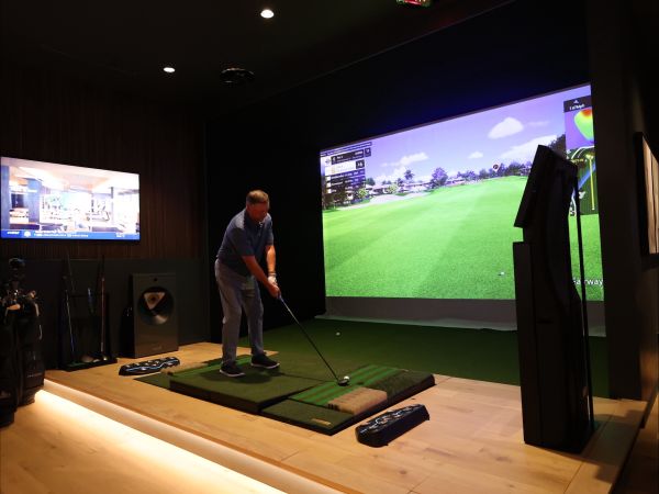A golfer mid-swing in an indoor simulator with performance data displayed on screens.